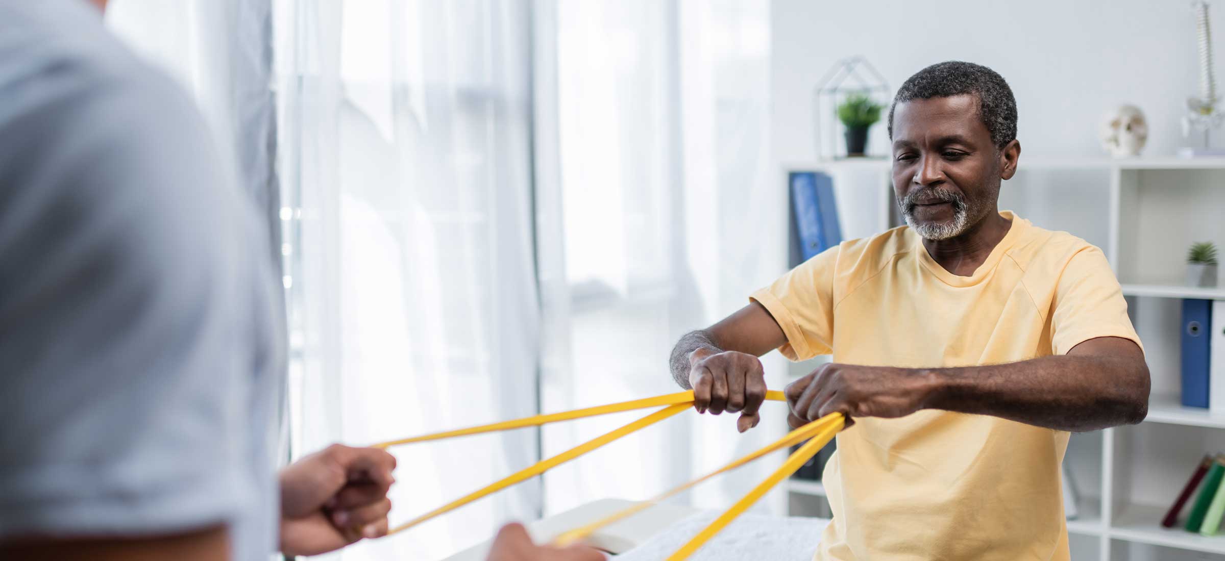 A physical therapist helps a senior man using exercise bands at a physical rehabilitation center.