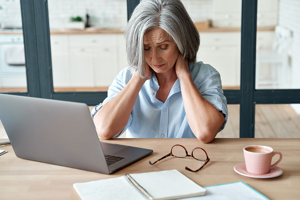 A middle-aged woman leans over her desk looking tired and touching her neck