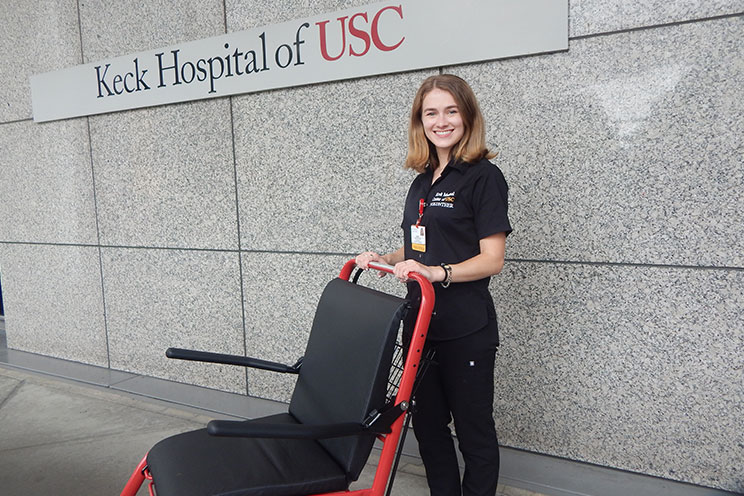 A cheerful Keck Medical Center of USC volunteer standing outside of a hospital building ready to assist with a wheelchair