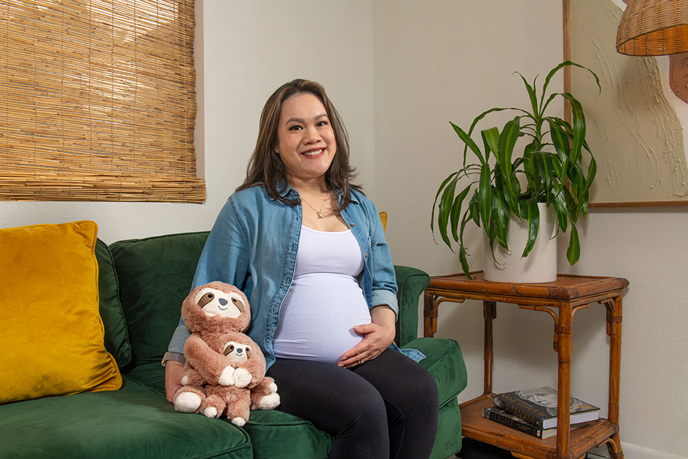Jocelyn Aguilar, a breast cancer survivor, sits on a green couch, holding a stuffed toy sloth with one arm and touching her pregnant belly with the other