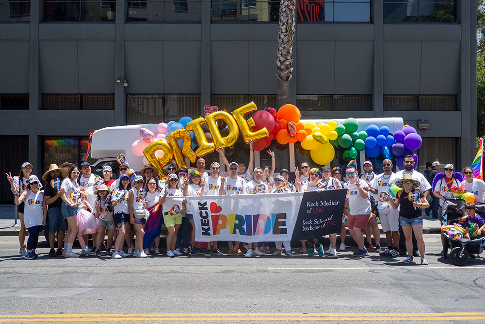 A diverse group of people standing in front of a colorful banner and holding rainbow-colored balloons for an outdoor parade.