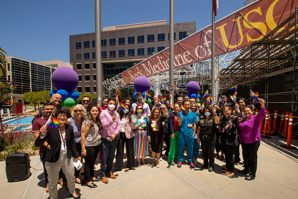 A diverse group of people standing in front of a colorful banner and holding rainbow-colored balloons for an outdoor parade.