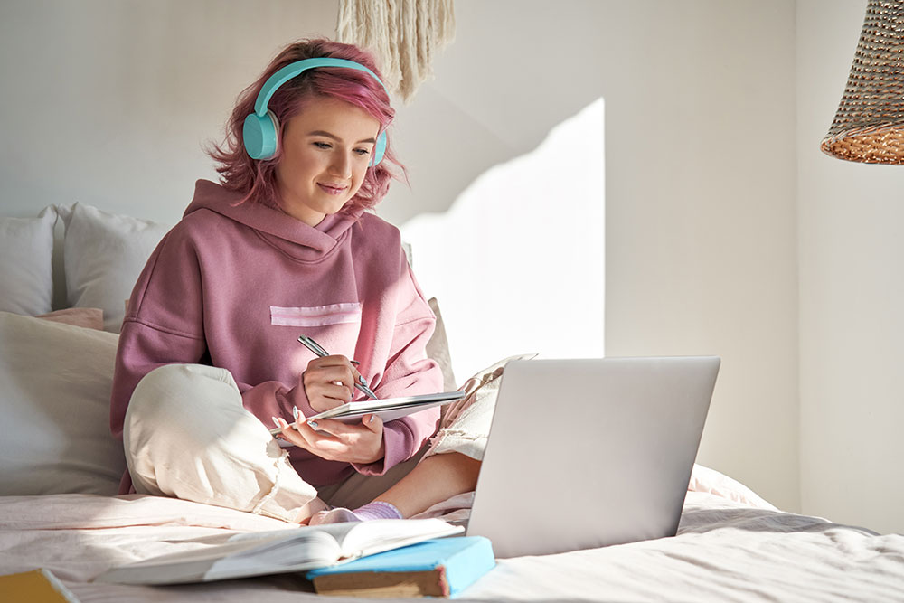 Hipster teen student with pink hair wears headphones and writes notes while watching a webinar in her bedroom at home.