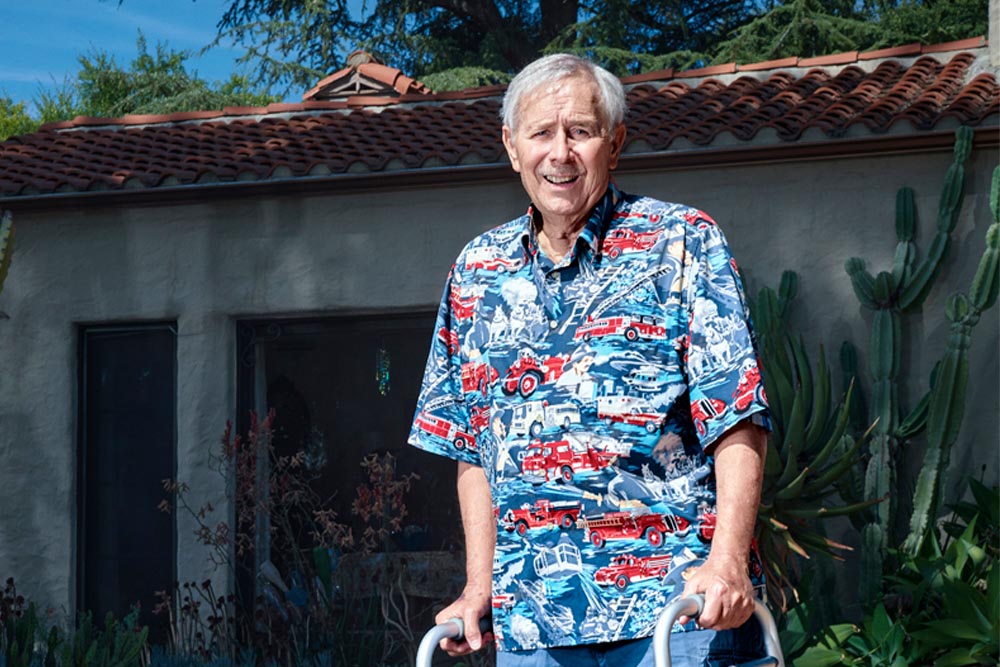 Gary Hood, an older caucasian man in a firefighting-themed shirt, stands outside his Sierra Madre home with a walker for support