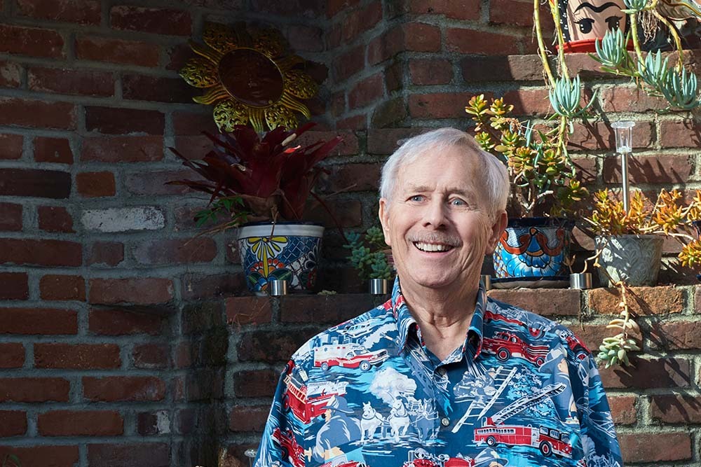 Heart attack patient Gary Hood, an older white man, smiles in his front yard, wearing a firefighting themed collared shirt