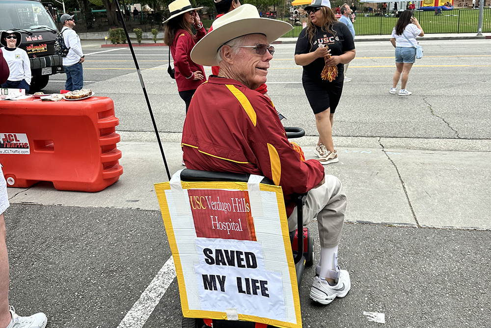Jim Lewis sits in a motorized scooter wearing a USC cardinal and gold fleece and wide-brimmed hat with a sign of the USC Verdugo Hills Hospital logo that says "USC Verdugo Hills Hospital saved my life"