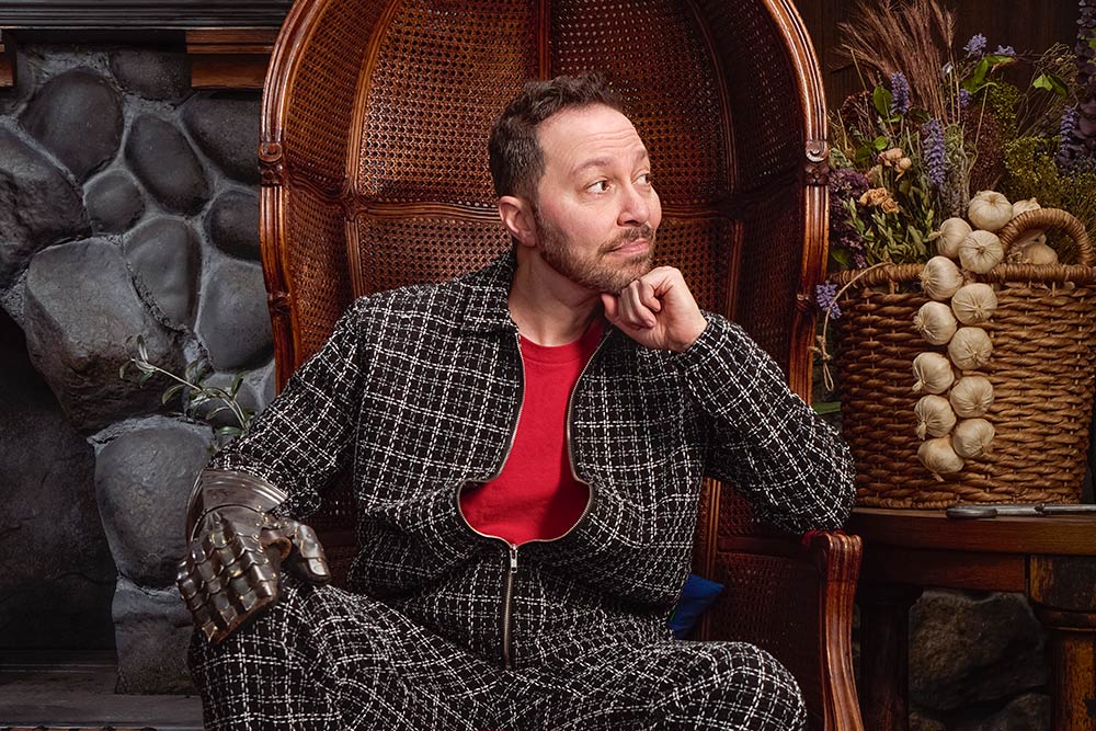 Sam Riegel sits in a high-backed chair with a suit of armor's glove over his hand