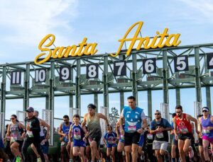 A large group of runners of various ages and backgrounds bursts forward from the starting gate of a horse racing track.