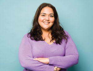A smiling woman with long brown hair, wearing a purple V-neck sweater, standing with her arms crossed against a light blue background.
