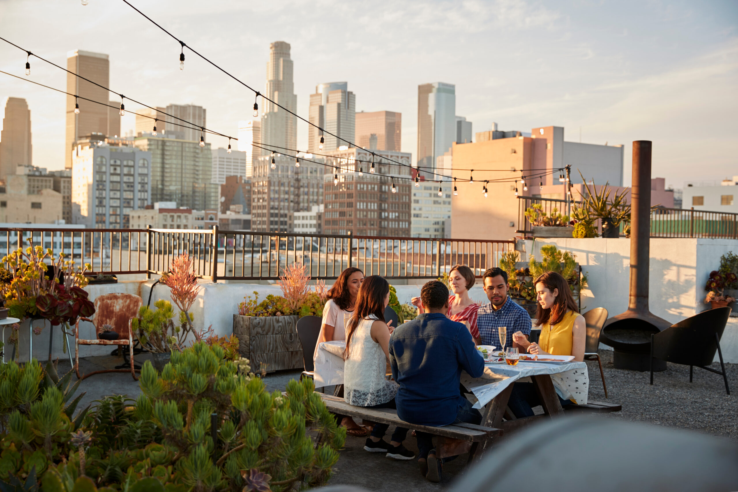 Friends Gathered On Rooftop Terrace For Meal With City Skyline In Background.