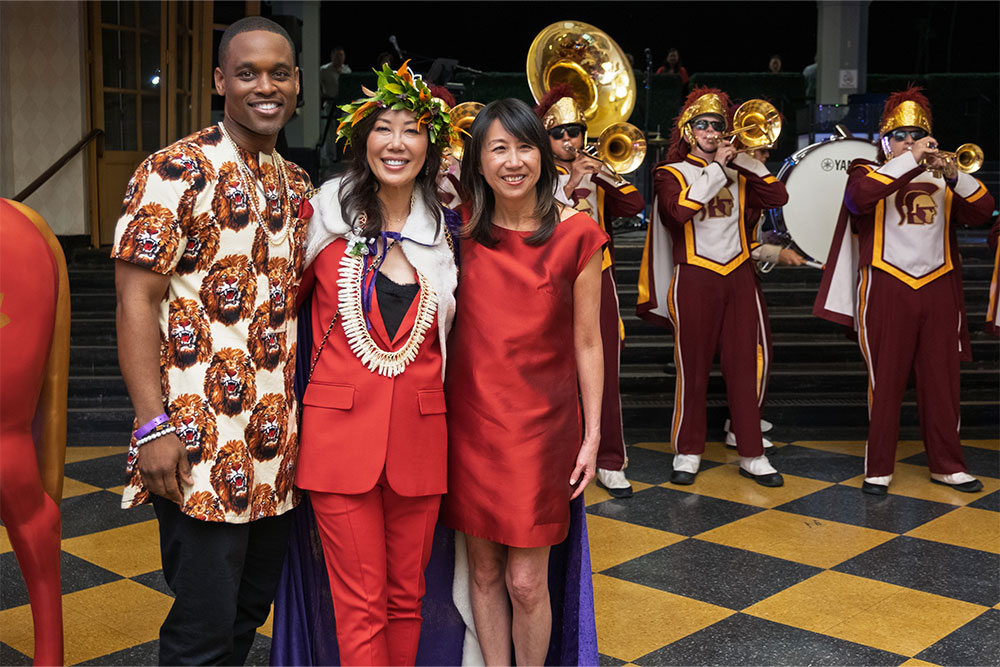 A smiling man and two women, dressed in red and one wearing a crown, in front of the USC Marching Band.