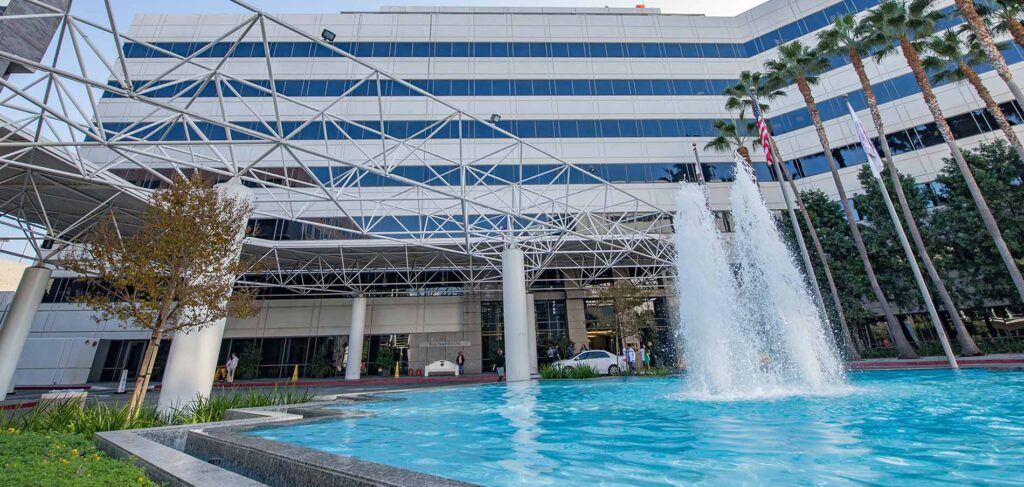 An exterior view of Keck Hospital of USC, a modern white multi-story building with a large decorative fountain spraying water into a bright blue pool in the foreground.