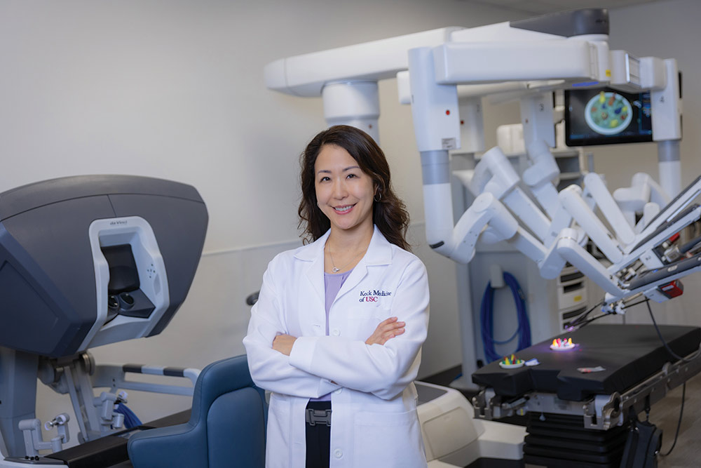 Sharon Shiraga in a white Keck Medicine lab coat stands in front of two surgical robots