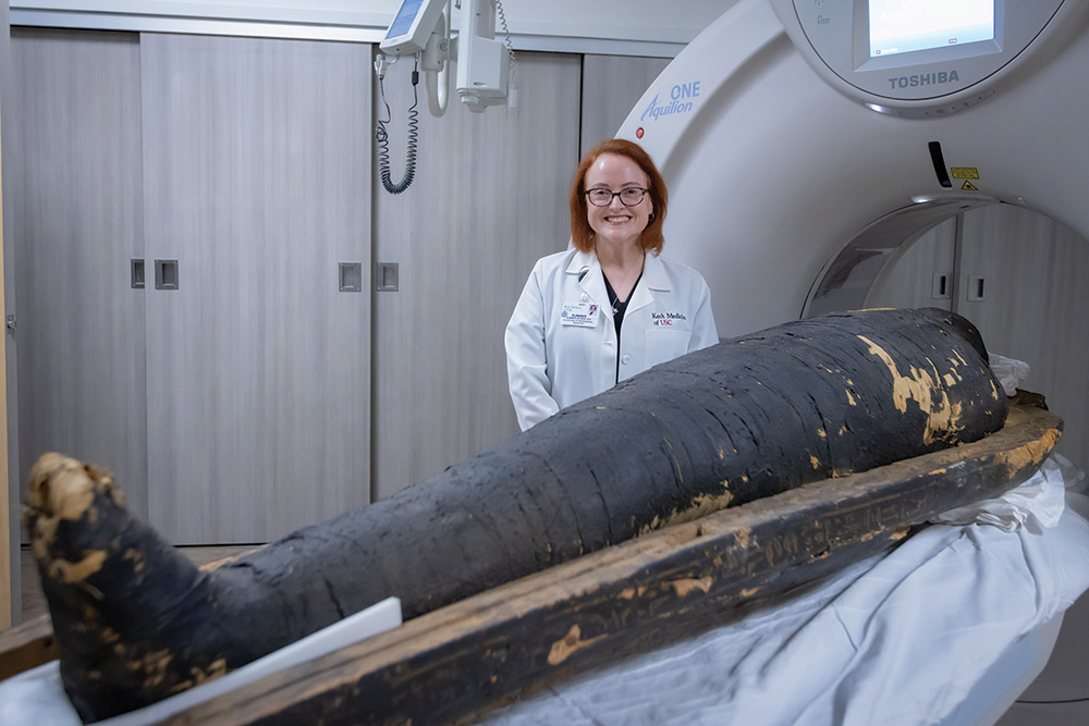 Dr. Summer Decker poses next to a CT scanner with an Egyptian mummy