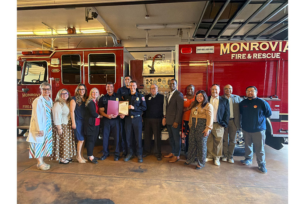 Women and men stand around three first responders in front of a fire truck.