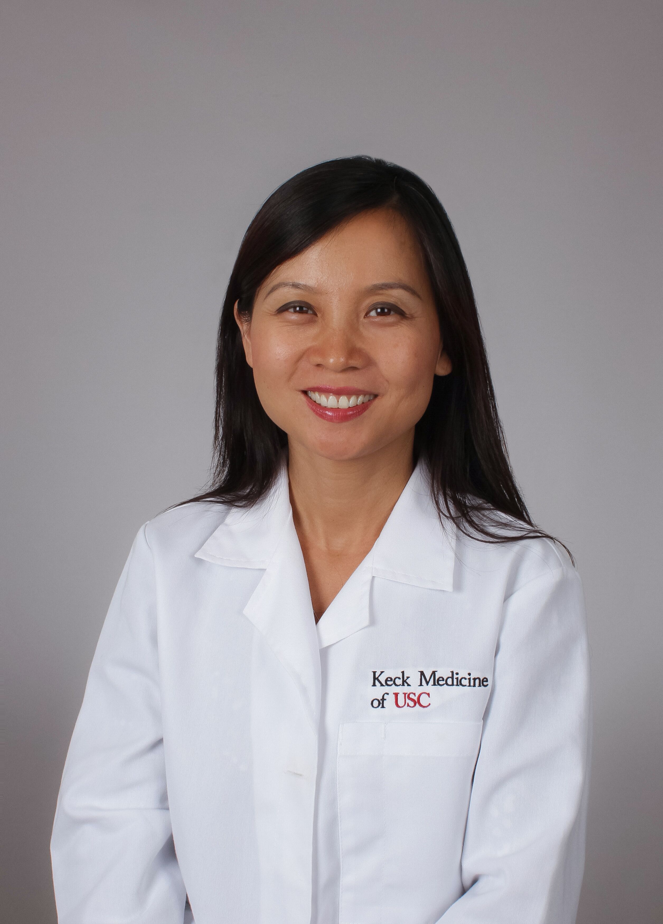 Dr. Liyun Yuan stands smiling in a white medical coat against a grey background