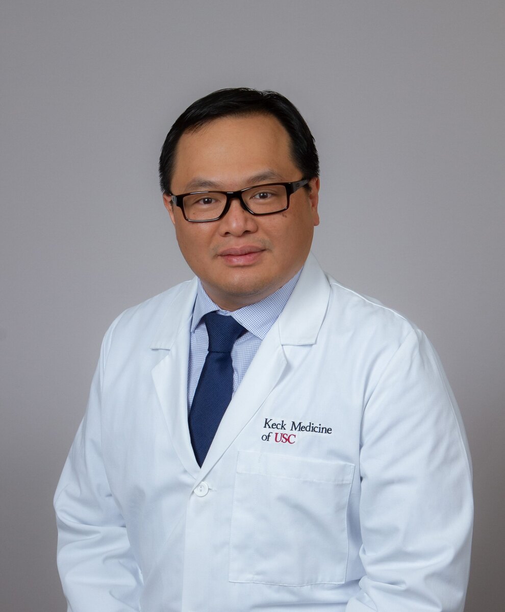 Dr. Ho stands in a white labcoat, smiling against a grey background