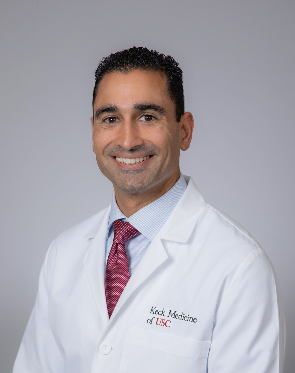 Dr. Malhotra stands smiling in a white medical coat against a grey background