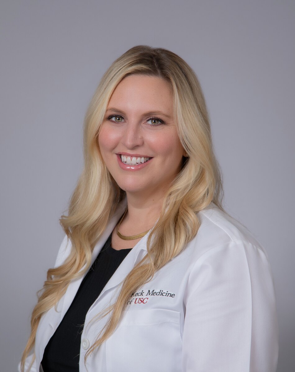 Dr. Shuman stands smiling in a white medical coat against a grey background